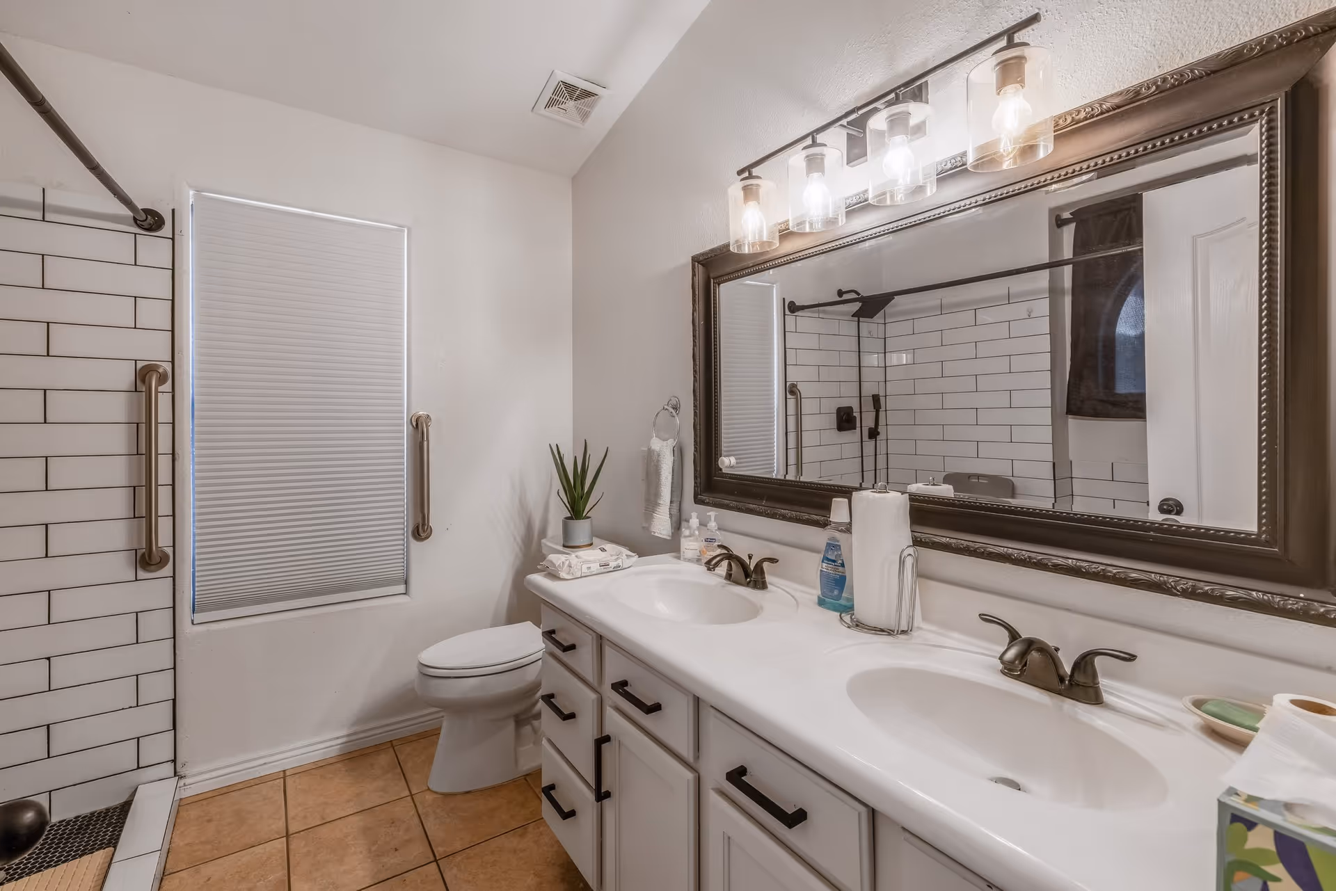 A bathroom with a double sink vanity featuring dark bronze faucets and a large framed mirror above. There is a toilet next to a window with a closed blind, and a shower area with white subway tiles and a dark bronze grab bar and shower fixtures. The floor is tiled in a warm brown color, and a small potted plant is placed on the vanity.