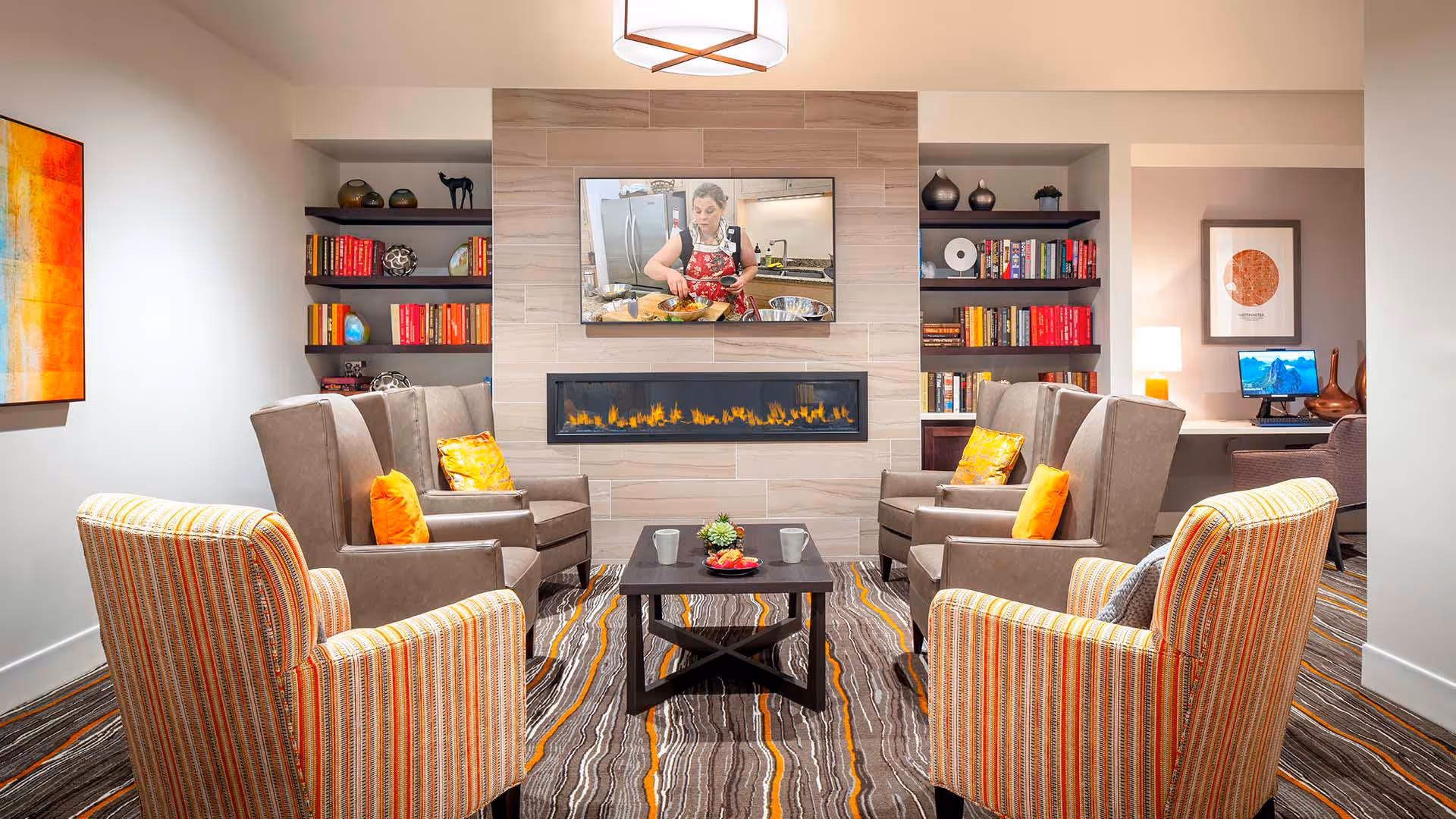Cozy communal lounge with armchairs arranged around a coffee table facing a wall-mounted TV above a linear fireplace and built-in bookshelves.