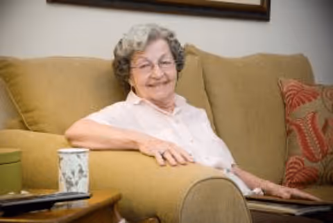 An elderly woman with gray curly hair and glasses sitting comfortably on a beige couch in a living room. She is smiling and wearing a light pink blouse. There is a wooden side table next to her with a decorative cup and a remote control on it. A red patterned pillow is visible on the couch.