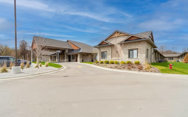 Front entrance of a single-story senior living facility with a covered drop-off drive, landscaped beds, and a blue sky.