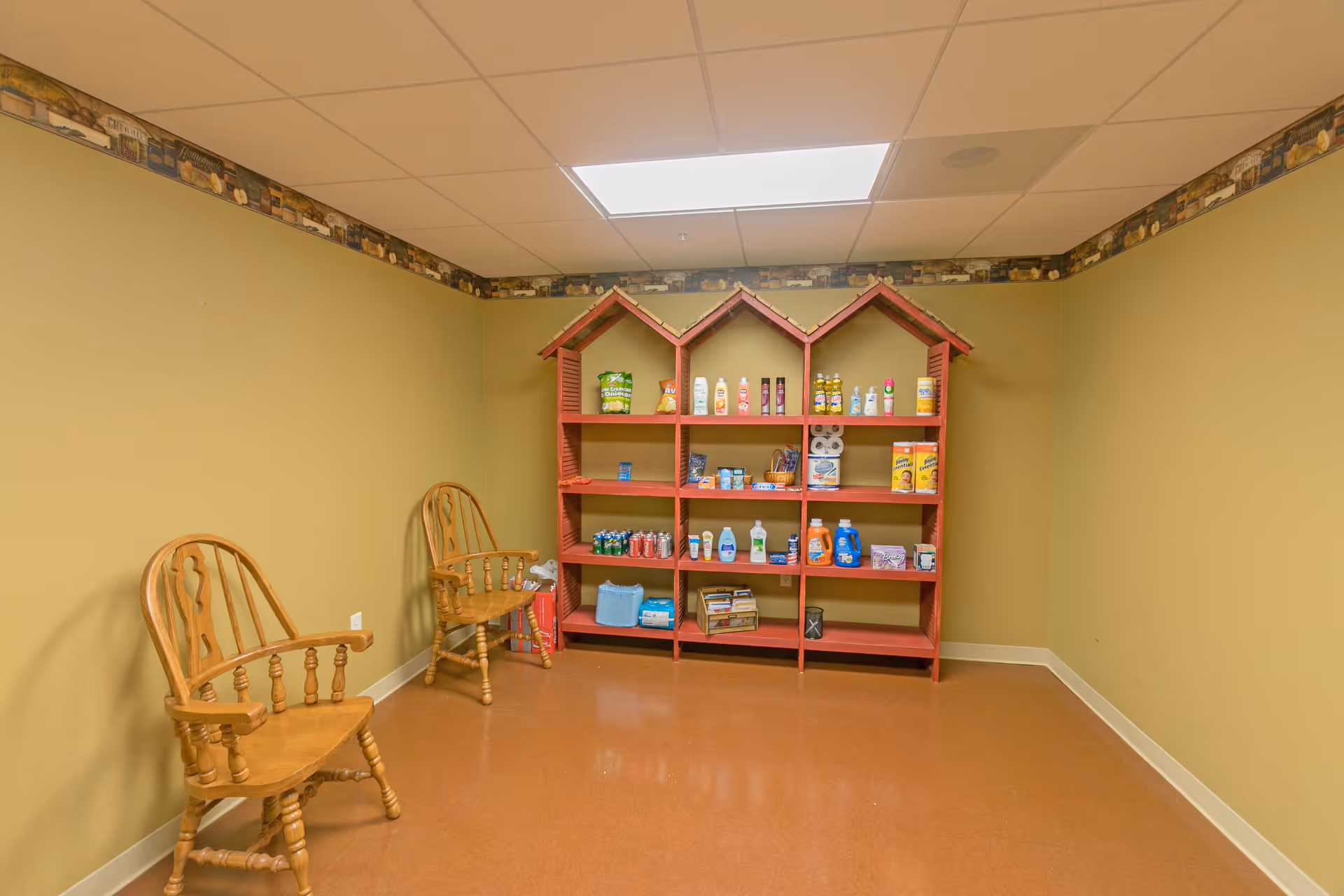 A small room with two wooden chairs and a red shelf shaped like three connected houses, stocked with various household items including cleaning supplies, canned drinks, and paper towels. The walls are painted beige with a decorative border near the ceiling, and the floor is a brownish color.