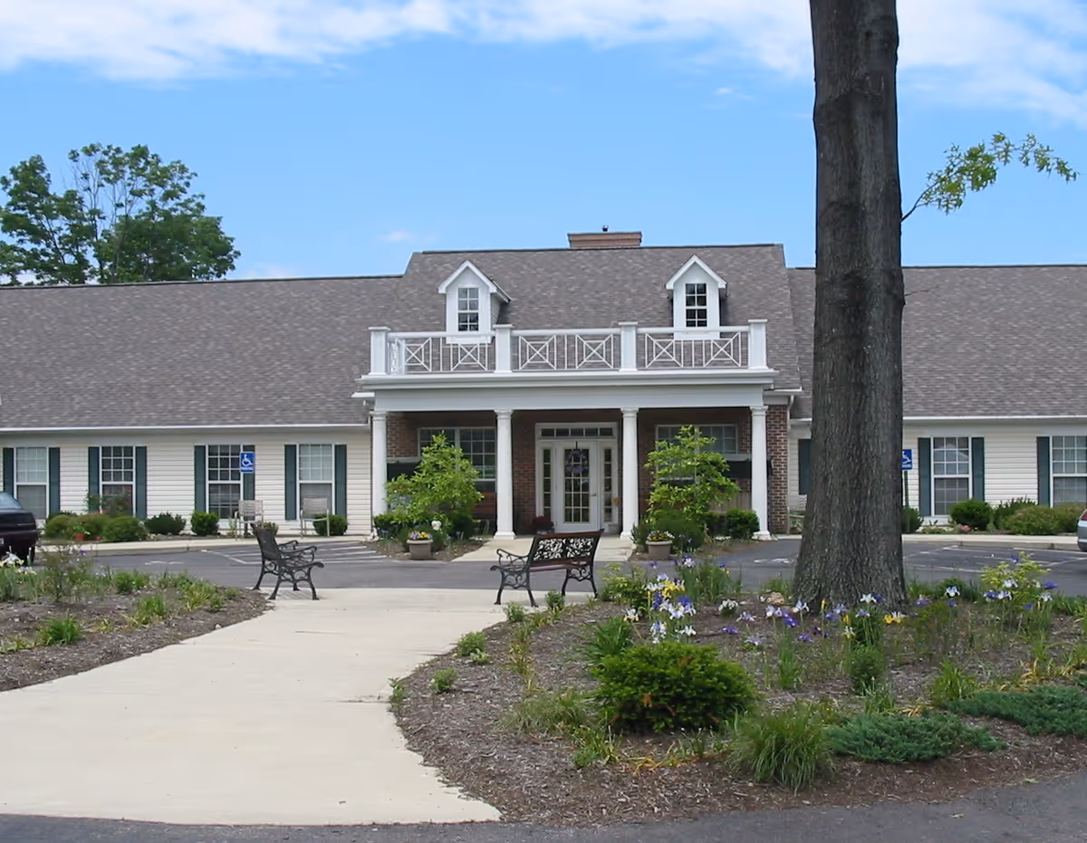 Front exterior view of Abbington of Powell Assisted Living building with a paved walkway leading to the entrance, benches on either side, landscaped garden beds with flowers and shrubs, and a large tree in the foreground under a partly cloudy sky.