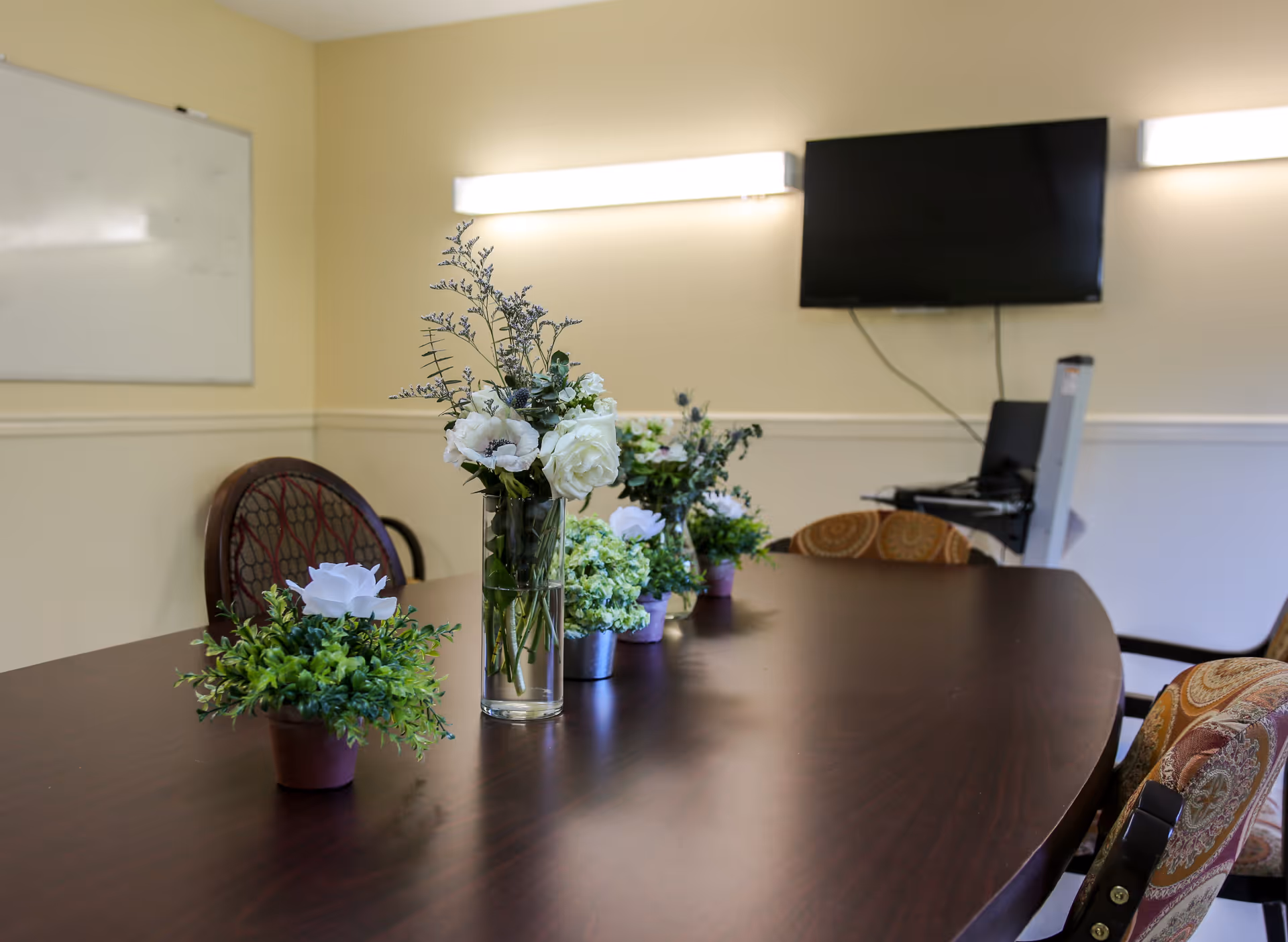 A conference or meeting room with a long wooden table adorned with several small flower arrangements. There are cushioned chairs around the table, a wall-mounted flat screen TV, two rectangular wall lights, and a whiteboard on the wall.