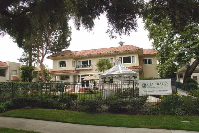 Front exterior of a two-story memory care building with a fenced garden, gazebo, and a 'Silverado' sign.