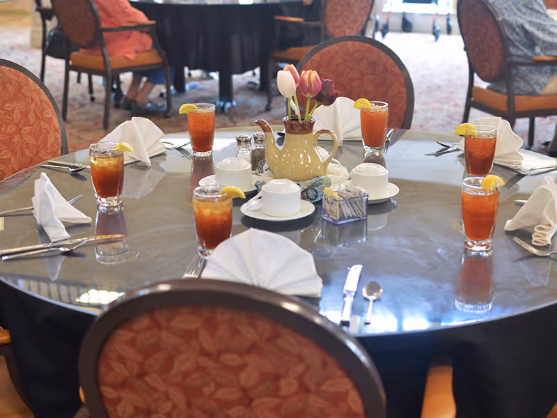 A round dining table set for six with glasses of iced tea garnished with lemon slices, white folded napkins, silverware, a ceramic pitcher with tulips, salt and pepper shakers, and sugar packets in a senior living facility dining room.