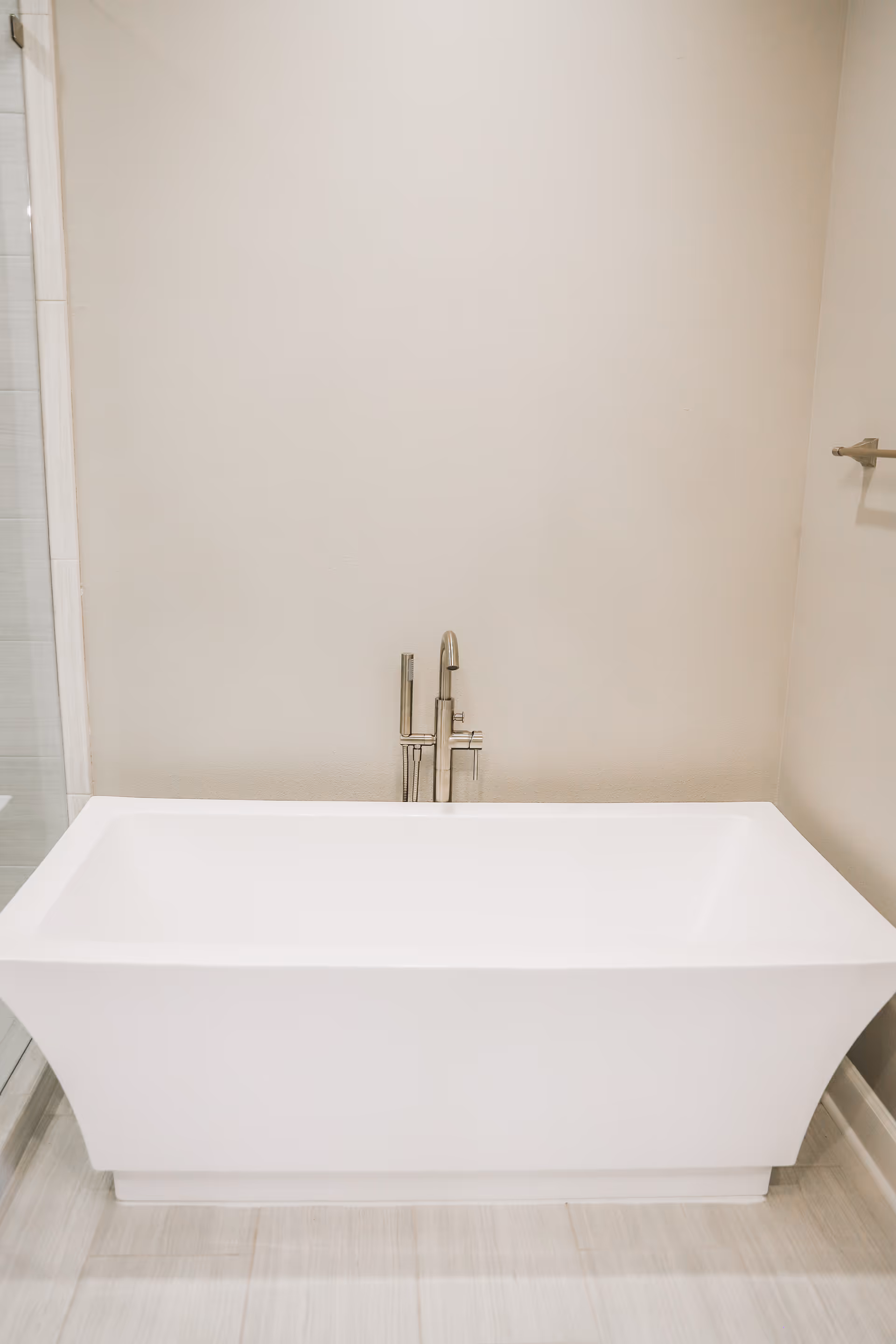 A modern freestanding white bathtub against a beige wall with a floor-mounted chrome faucet.