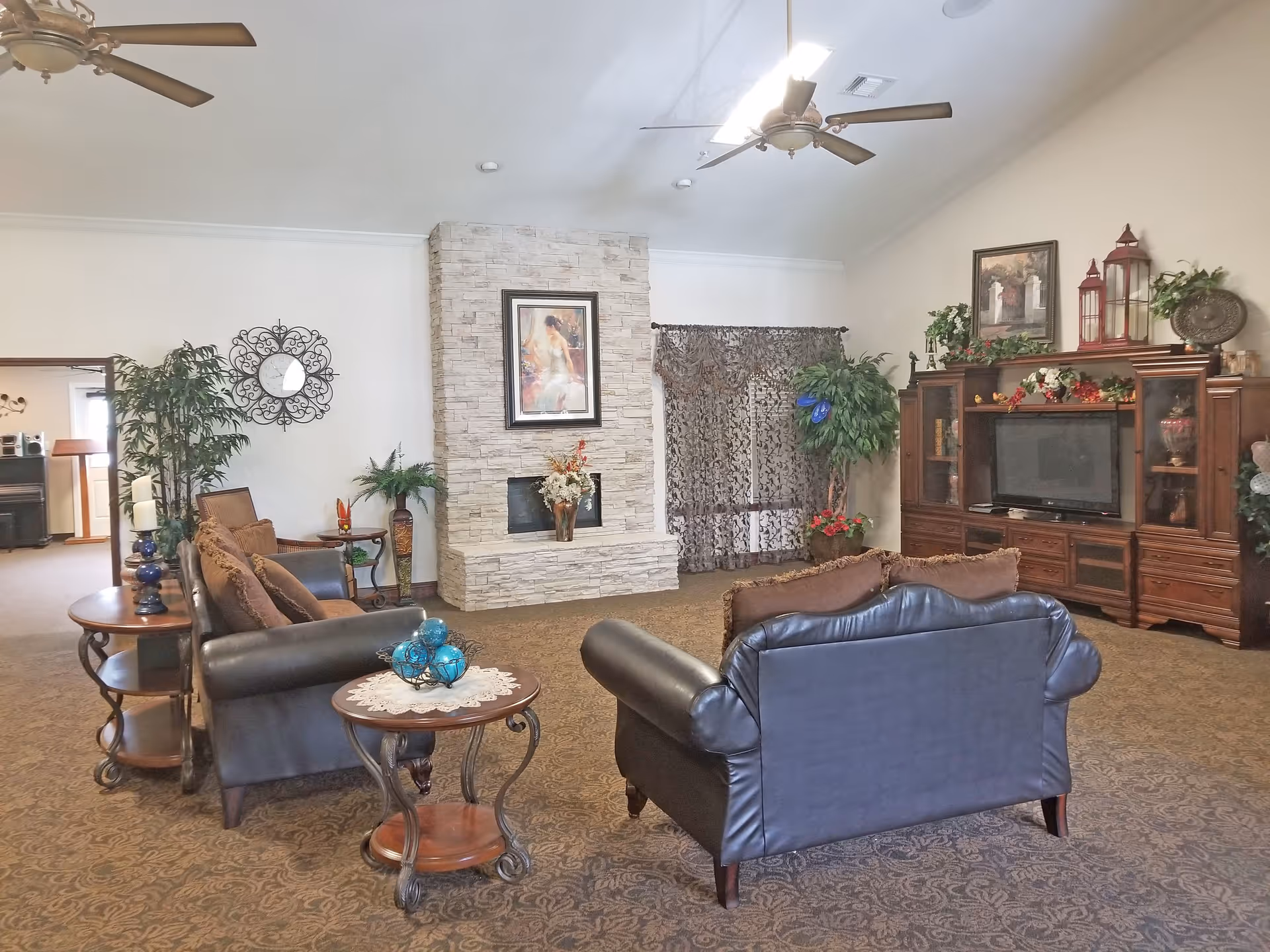Spacious furnished common living room with leather sofas, a stone fireplace, TV cabinet, and decorative plants.