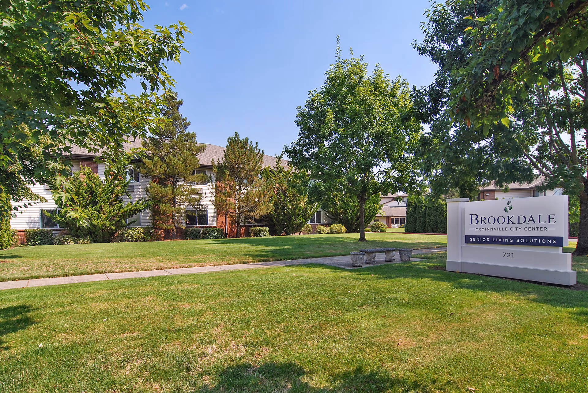 Exterior view of Brookdale McMinnville City Center senior living facility with a well-maintained lawn, trees, and a sidewalk. A white sign with the facility name and address 721 is visible on the right side of the image.