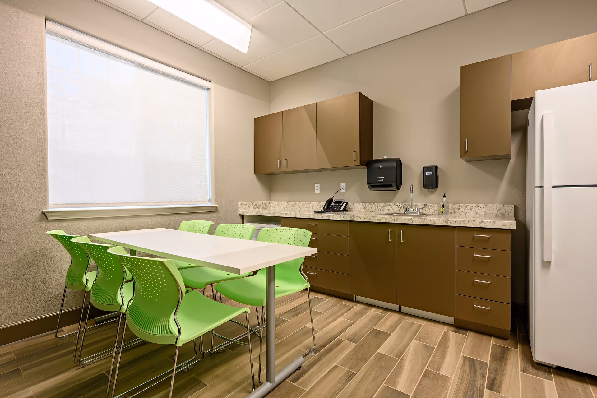 A small kitchen area with brown cabinets, a white refrigerator, a countertop with a sink, a telephone, a paper towel dispenser, and a soap dispenser. In front of the kitchen counter is a white rectangular table surrounded by six green chairs. The room has wood-patterned tile flooring and a window with a white roller shade.
