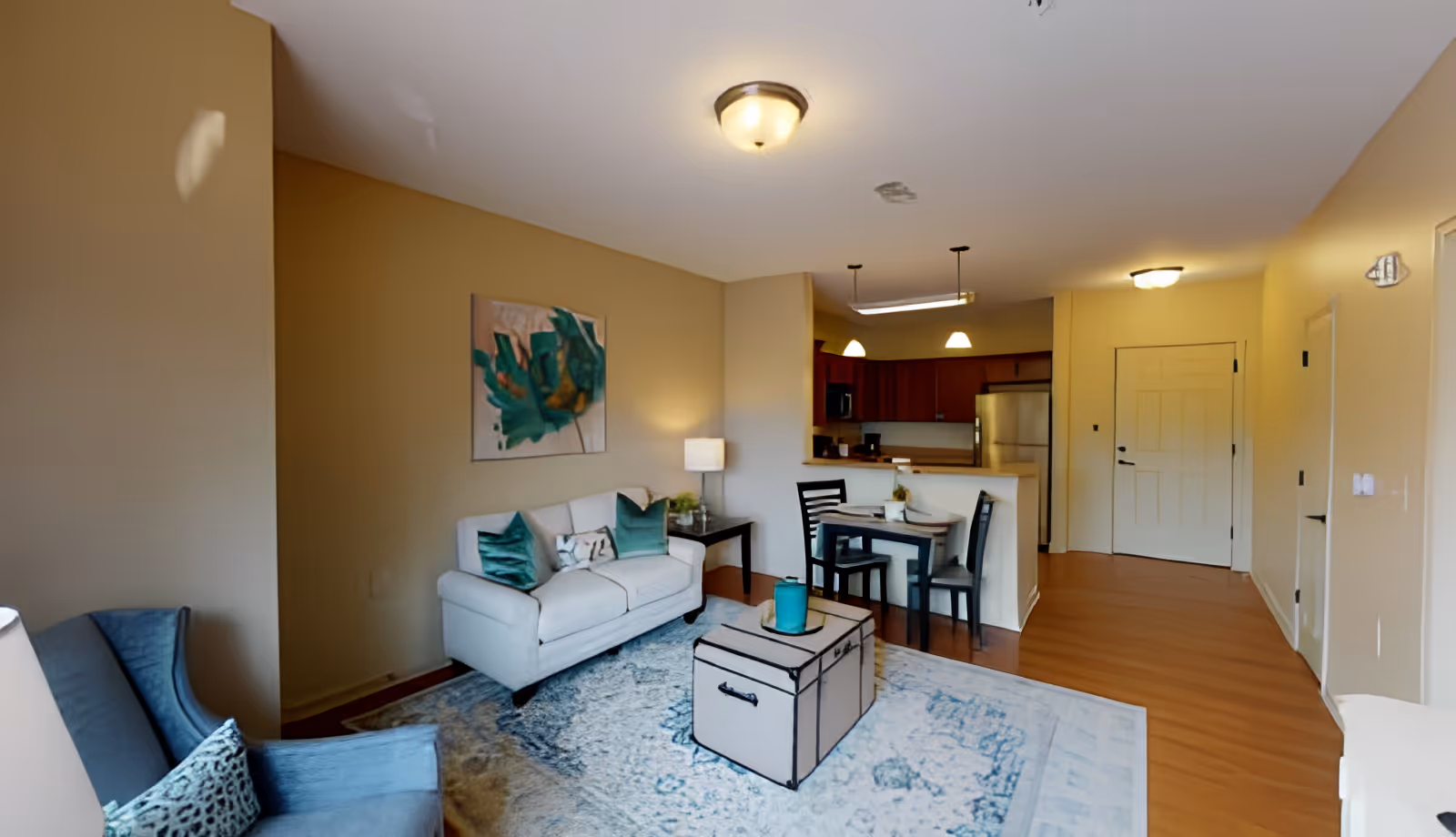 A cozy living room area with a white loveseat adorned with teal and patterned pillows, a blue armchair with a patterned pillow, a beige trunk-style coffee table on a blue and white patterned rug, and a side table with a lamp. The room opens into a small dining area with a table and four chairs, and a kitchen with wooden cabinets and stainless steel appliances. The walls are painted a warm beige color, and there is a piece of abstract art hanging above the loveseat.