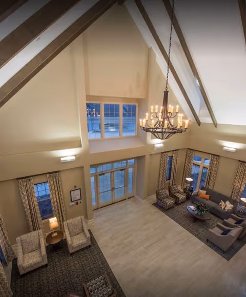 Spacious living room area with high vaulted ceilings and exposed wooden beams. The room features a large chandelier hanging from the ceiling, multiple windows with patterned curtains, and a seating arrangement including armchairs, a sofa, and side tables with lamps. The floor is a combination of carpet and light-colored tile.