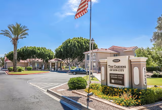 Exterior view of The Gardens of Sun City assisted living facility with a clear blue sky, an American flag on a flagpole, landscaped bushes and trees, a driveway, and a sign displaying the facility's name and services.