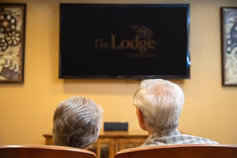 Two elderly individuals sitting side by side in chairs, facing a television mounted on a wall displaying the text 'The Lodge at Natchez Trace'. The room has warm-colored walls and framed artwork on either side of the television.