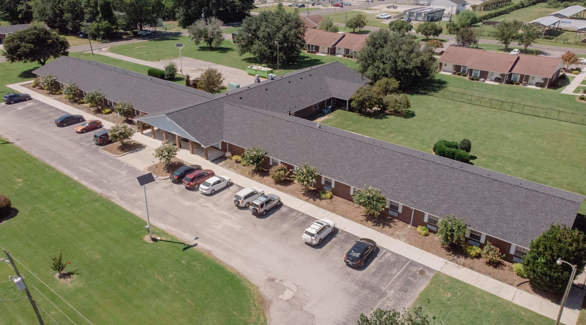 Aerial view of a single-story L-shaped brick building with a covered entrance, parked cars, and surrounding grassy grounds.