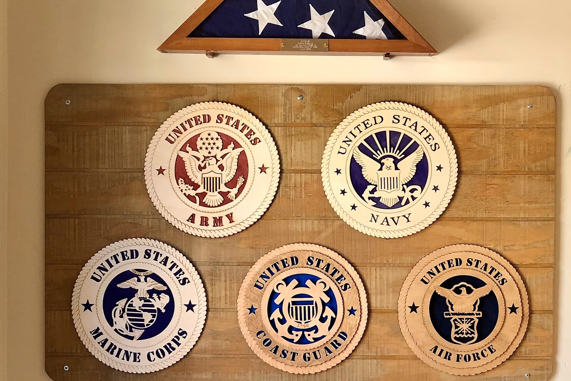 Wooden plaques displaying the emblems of five branches of the United States military: Army, Navy, Marine Corps, Coast Guard, and Air Force, mounted on a wooden board with a folded American flag in a triangular display case above them.