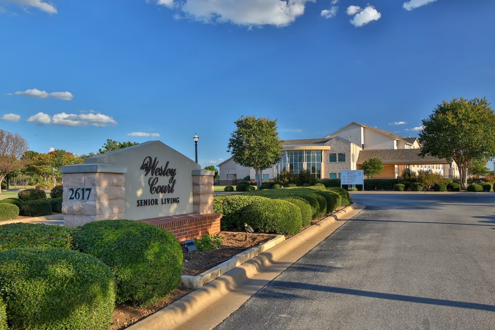 Exterior view of Wesley Court Senior Living facility with a clear blue sky, manicured bushes, trees, and a paved driveway leading to the building entrance. A large sign with the facility name and address number 2617 is prominently displayed in the foreground.