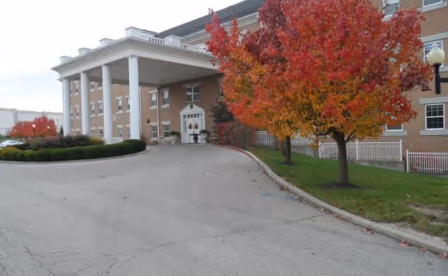 Entrance of a multi-story brick building with large white columns supporting a covered driveway. Trees with vibrant red and orange autumn leaves line the driveway on the right side.