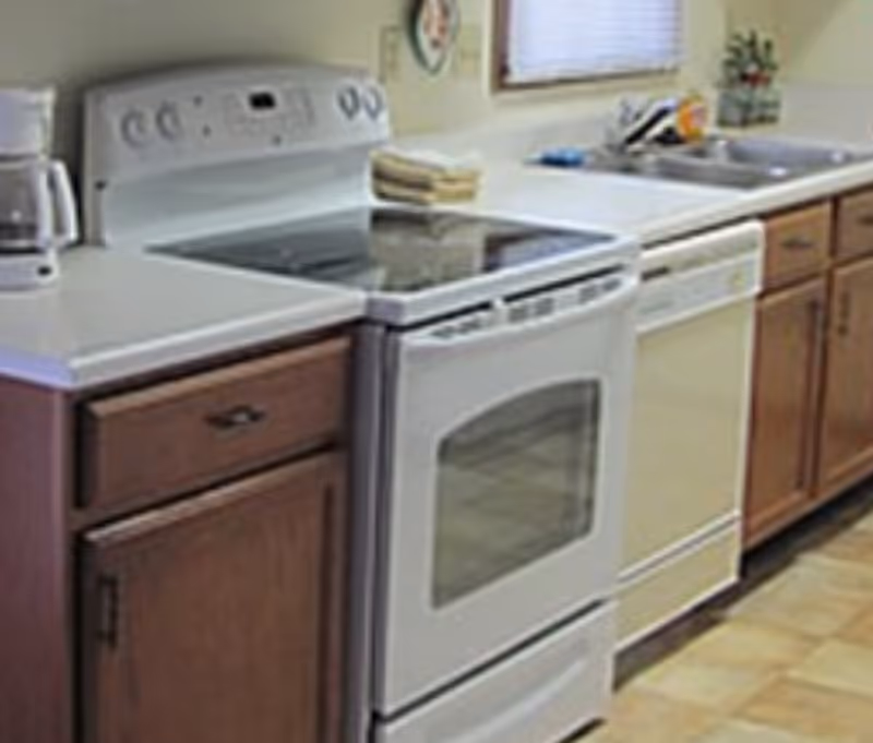 A kitchen area with a white electric stove, dishwasher, wooden cabinets, a coffee maker, and a double sink under a window with blinds.