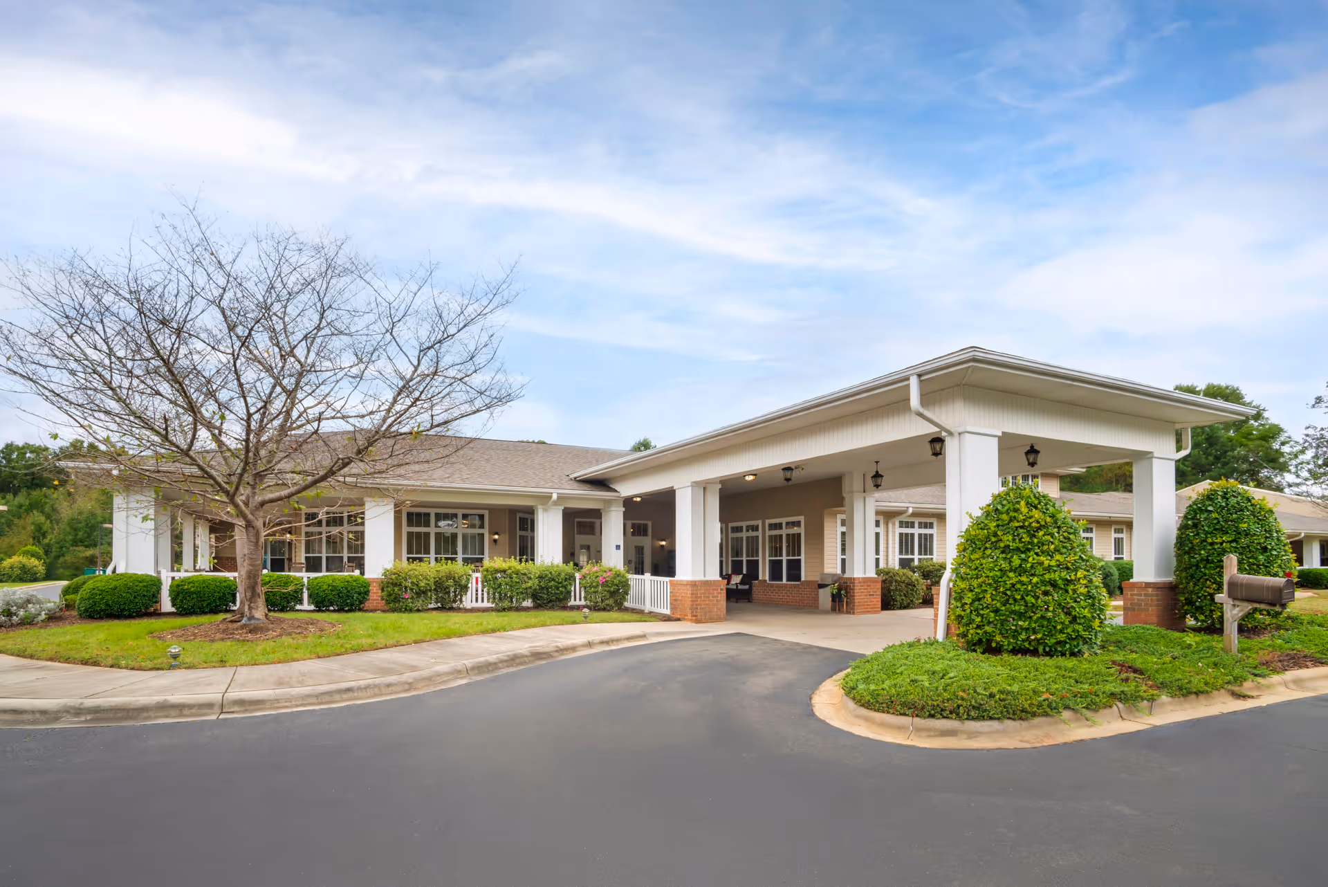 Exterior view of a single-story building with a covered entrance supported by white columns. The building is surrounded by neatly trimmed bushes, a leafless tree, and a well-maintained lawn. The sky is partly cloudy.