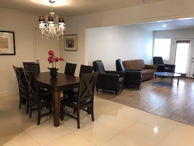 A combined dining and living room area with a dark wooden dining table and six matching chairs on a tiled floor. A chandelier with five lampshades hangs above the table, which has a vase with pink flowers. The living area has a brown sofa, two black armchairs, and a round coffee table on a wooden floor. There is a door and a window with blinds in the background.