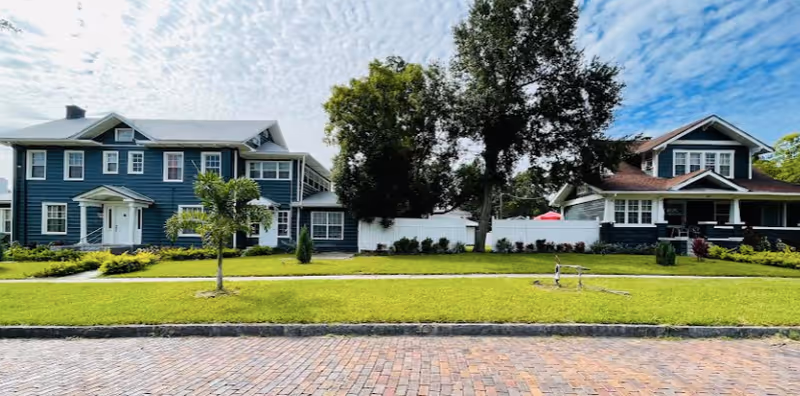 Two large residential-style buildings with multiple windows and porches, separated by a white fence and surrounded by green lawns and trees under a partly cloudy blue sky.