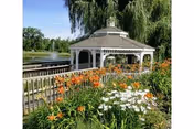 White gazebo beside a pond surrounded by orange and white flowers and willow trees.