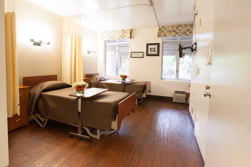 A bright room with two hospital-style beds covered with brown blankets, each with a small overbed table holding a vase of flowers. The room has wooden flooring, two windows with patterned valances, wall-mounted lights, and a small wooden nightstand between the beds. A chair is partially visible near the window.