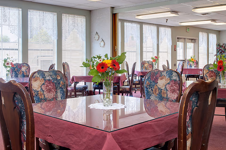 Dining room with glass-topped tables covered in burgundy tablecloths, floral-upholstered chairs, and vases of colorful flowers by large windows.