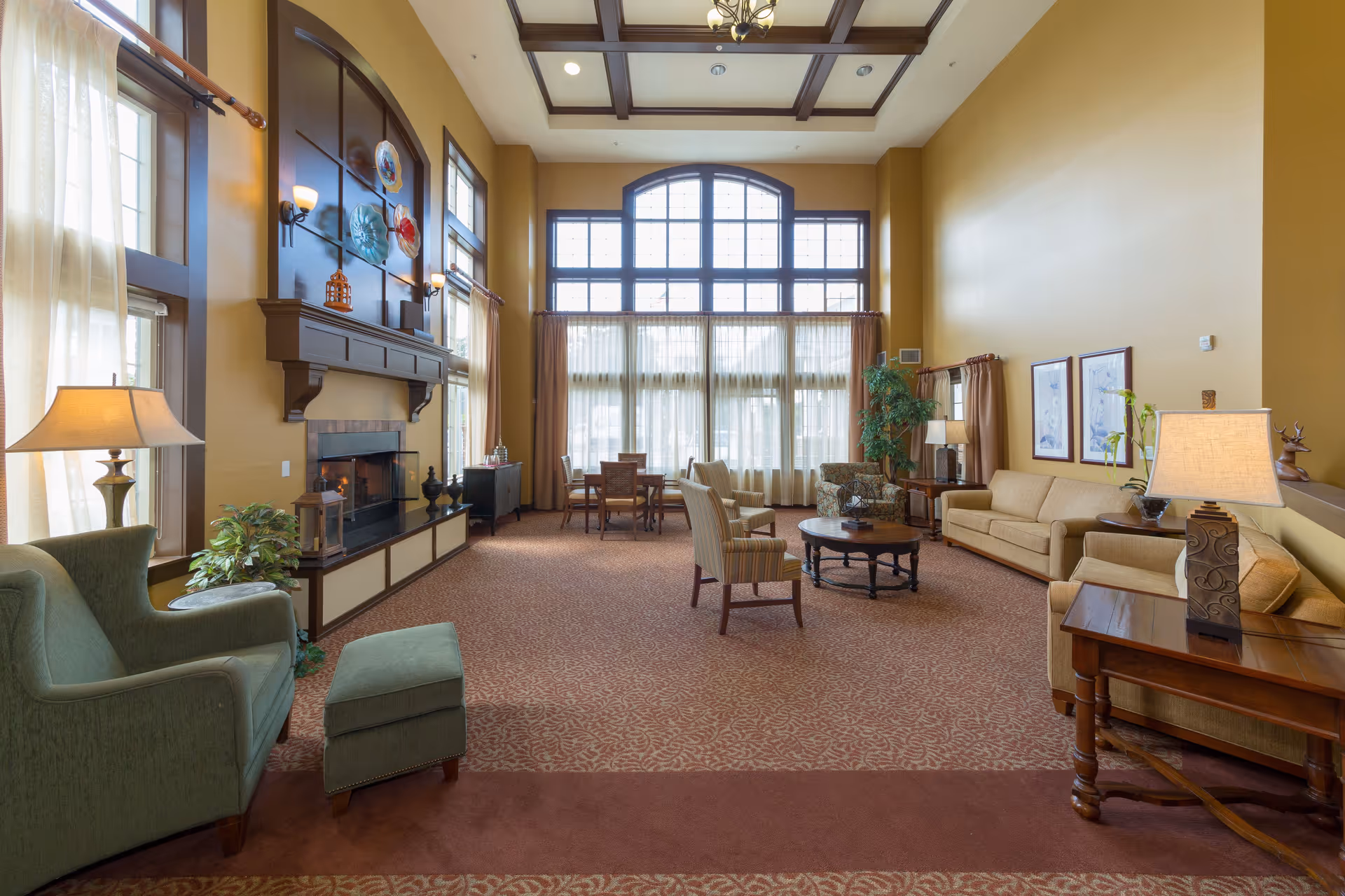 A spacious living room with high ceilings and large windows letting in natural light. The room features a fireplace with decorative plates above it, several armchairs, sofas, side tables with lamps, a coffee table, and a dining table with chairs near the windows. The walls are painted a warm beige color and the carpet has a patterned design.