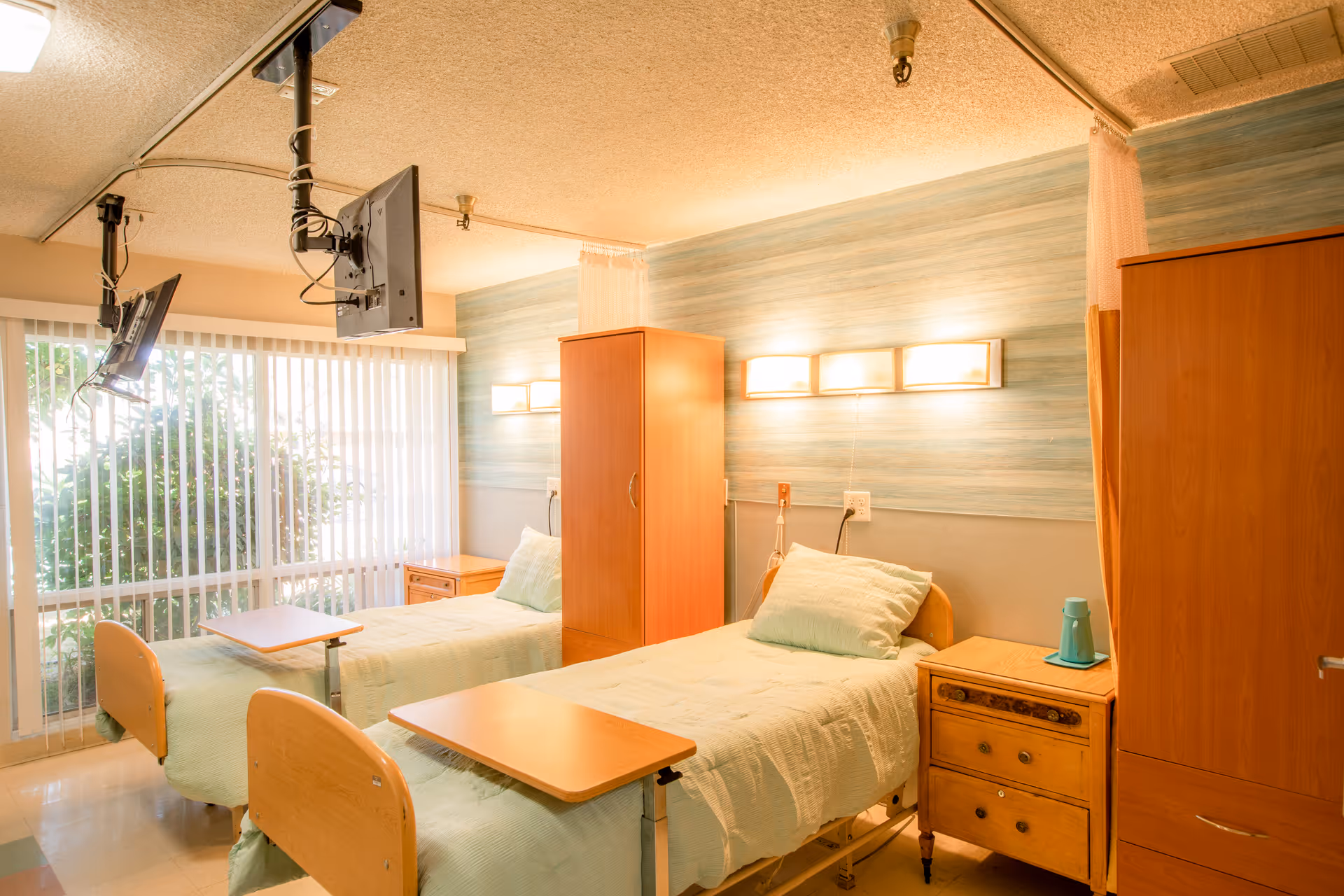 A bright and clean shared bedroom in a healthcare facility with two hospital beds, each with a light green bedspread and pillow. Each bed has an adjustable overbed table. There are wooden wardrobes and nightstands between and beside the beds. Two wall-mounted televisions hang from the ceiling. Large vertical blinds cover a window letting in natural light.