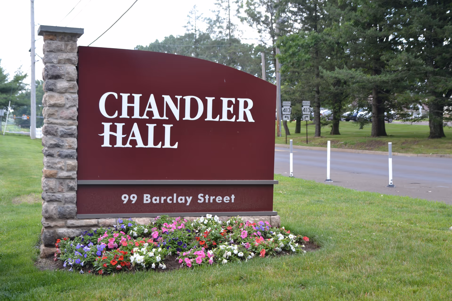 A large maroon sign with white text reading 'CHANDLER HALL' and '99 Barclay Street' is displayed on a stone base surrounded by colorful flowers. The sign is positioned on a grassy area next to a road with trees and traffic signs in the background.