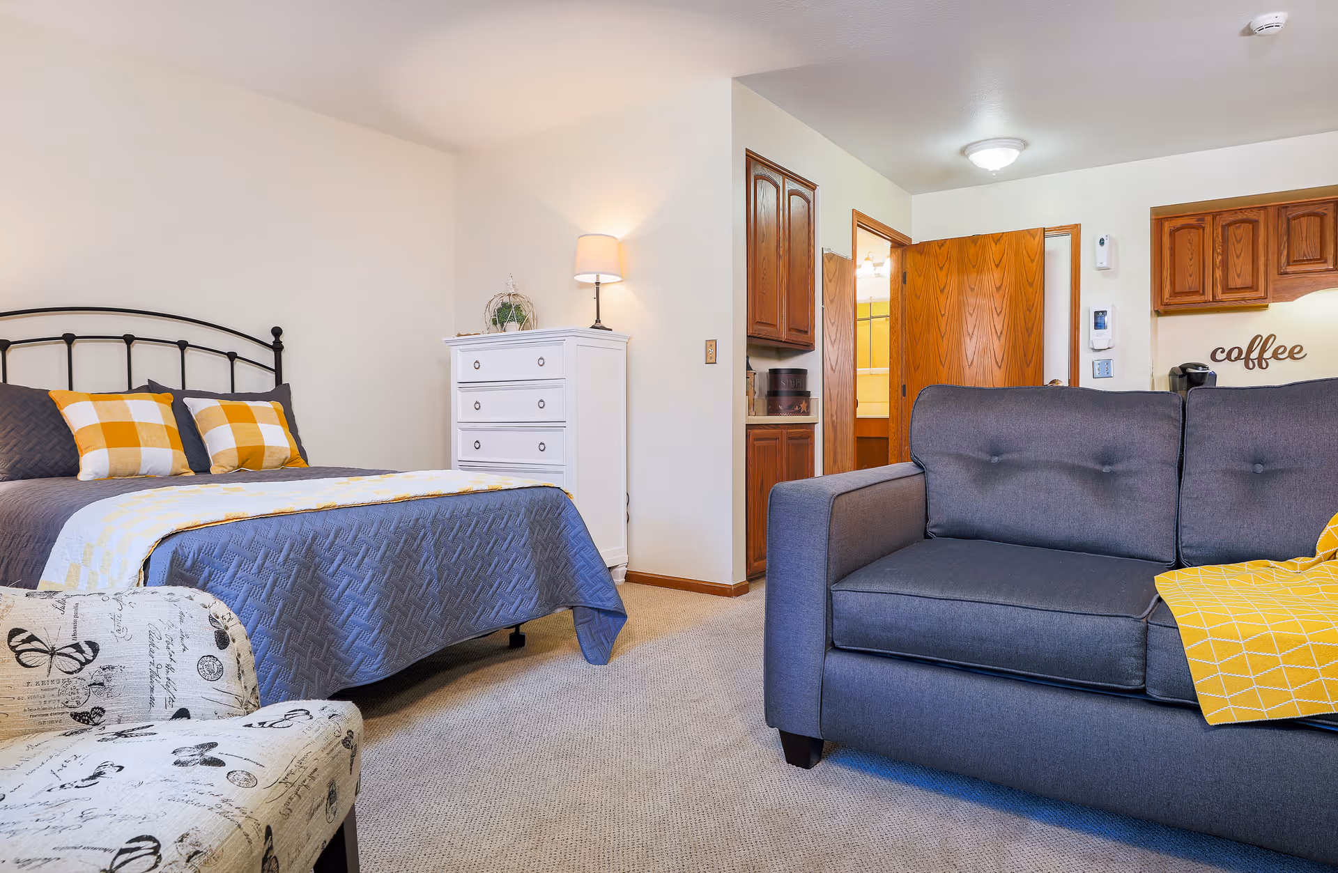 A cozy assisted living facility room featuring a bed with gray and yellow checkered pillows and a gray quilt, a white chest of drawers with a lamp and decorative plant on top, a gray sofa with a yellow throw blanket, a patterned armchair, and a small kitchenette area with wooden cabinets and a coffee station.