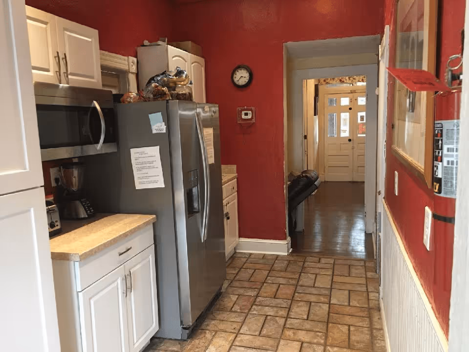 A kitchen area with white cabinets, a stainless steel refrigerator, microwave, and blender. The walls are painted red, and there is a tiled floor. A clock and thermostat are mounted on the wall, and a hallway with a door at the end is visible beyond the kitchen.