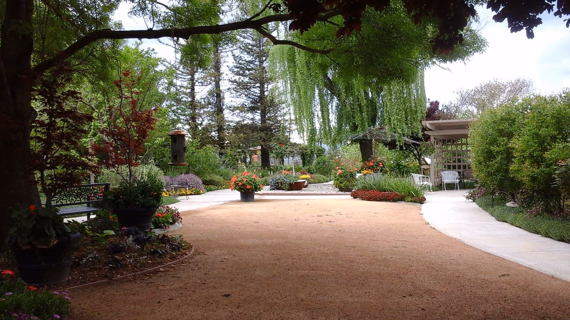 A peaceful garden area with a dirt pathway surrounded by lush greenery, colorful flowers, and trees. There are benches and a small gazebo with chairs on the right side, providing a relaxing outdoor space.
