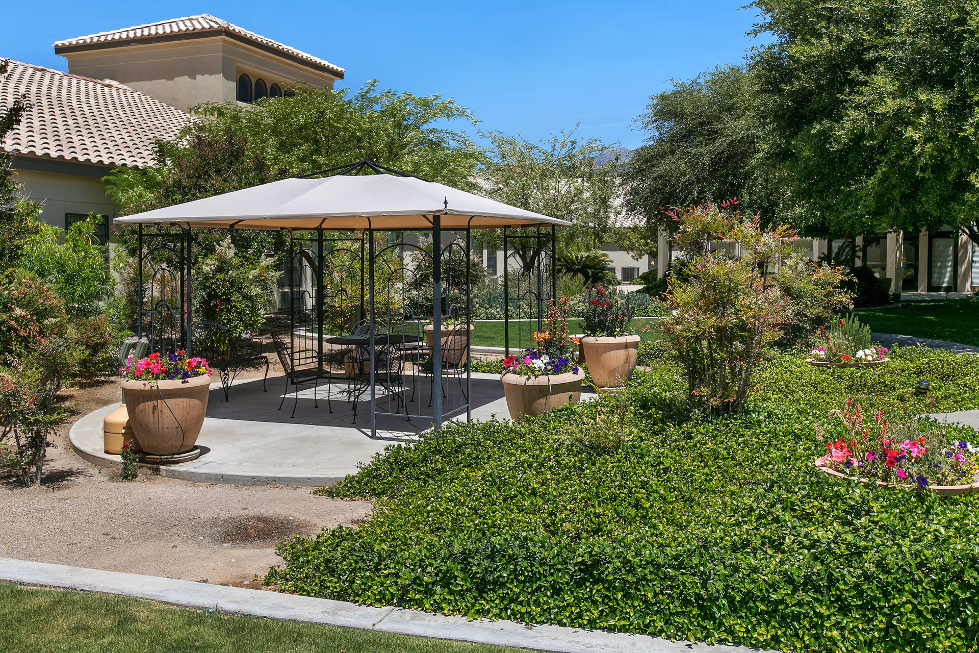Outdoor garden area at Inspirations of River Centre featuring a gazebo with a white canopy, metal chairs and tables underneath, surrounded by large flower pots with colorful flowers, green shrubs, and trees, with a building with a tiled roof in the background under a clear blue sky.