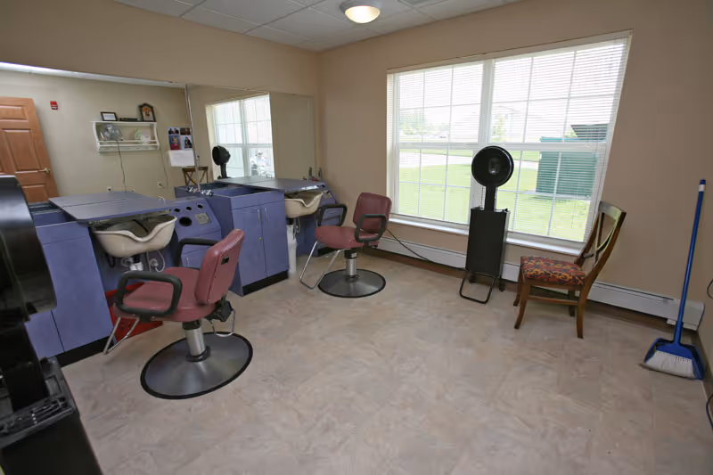Interior room with two salon chairs in front of sinks and mirrors, a window with blinds, a standing fan, a wooden chair with a patterned cushion, and a broom in the corner.