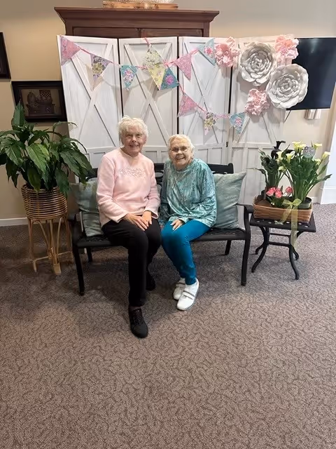 Two elderly women sitting on a black bench with cushions in a cozy indoor setting. Behind them is a decorative white folding screen adorned with pastel-colored triangular bunting and large white and pink paper flowers. To the left, there is a large green potted plant on a stand, and to the right, a small table with a flower arrangement. The floor is carpeted and a TV is mounted on the wall in the background.