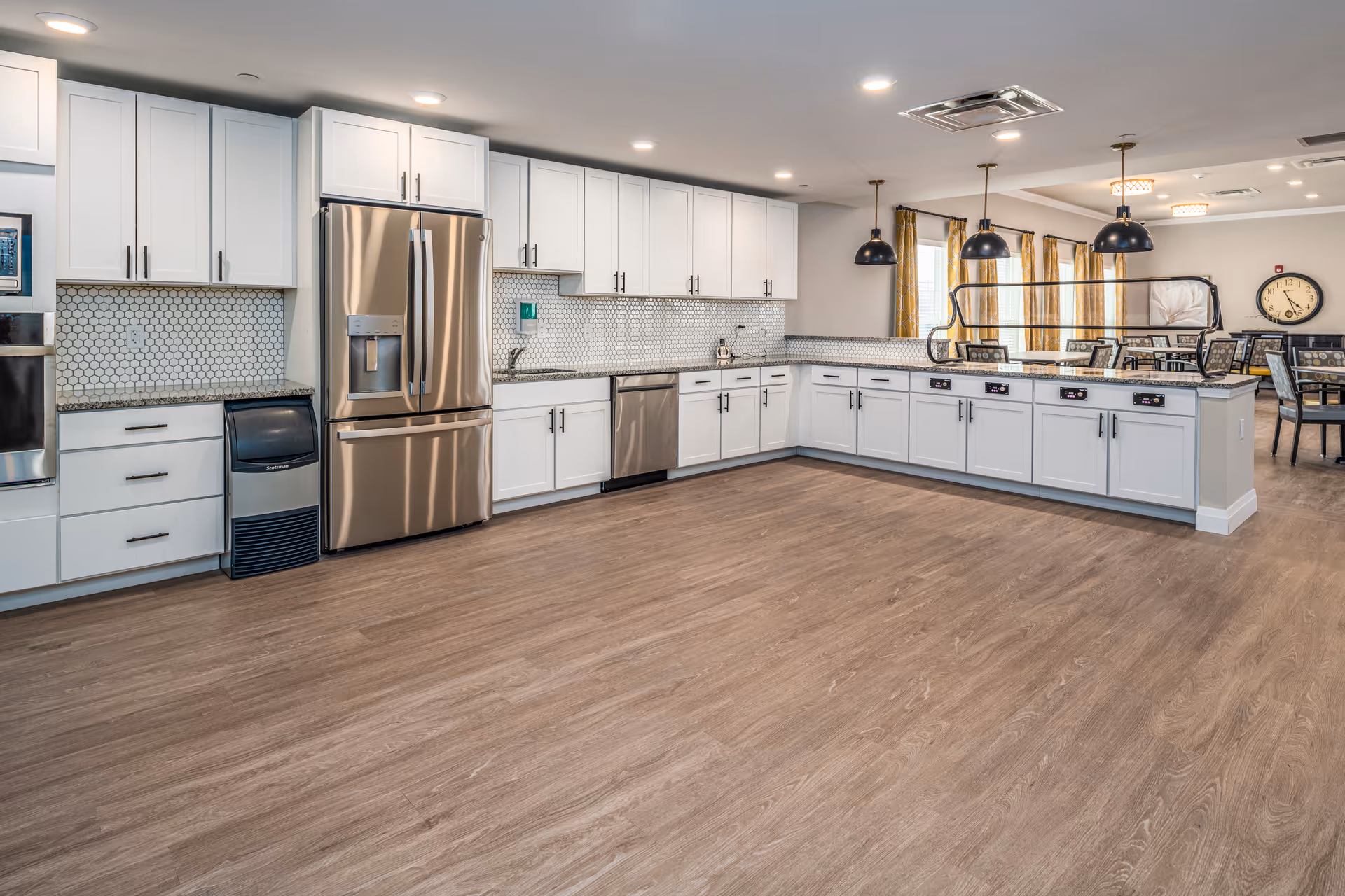 Large communal kitchen with white cabinets, stainless steel appliances, a long counter island and dining area in the background.