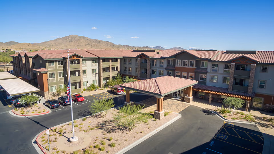 Exterior view of a senior living facility with a three-story building featuring a covered entrance, multiple windows, and a parking area with several cars. The landscape includes desert plants and a flagpole with an American flag. Mountains are visible in the background under a clear blue sky.