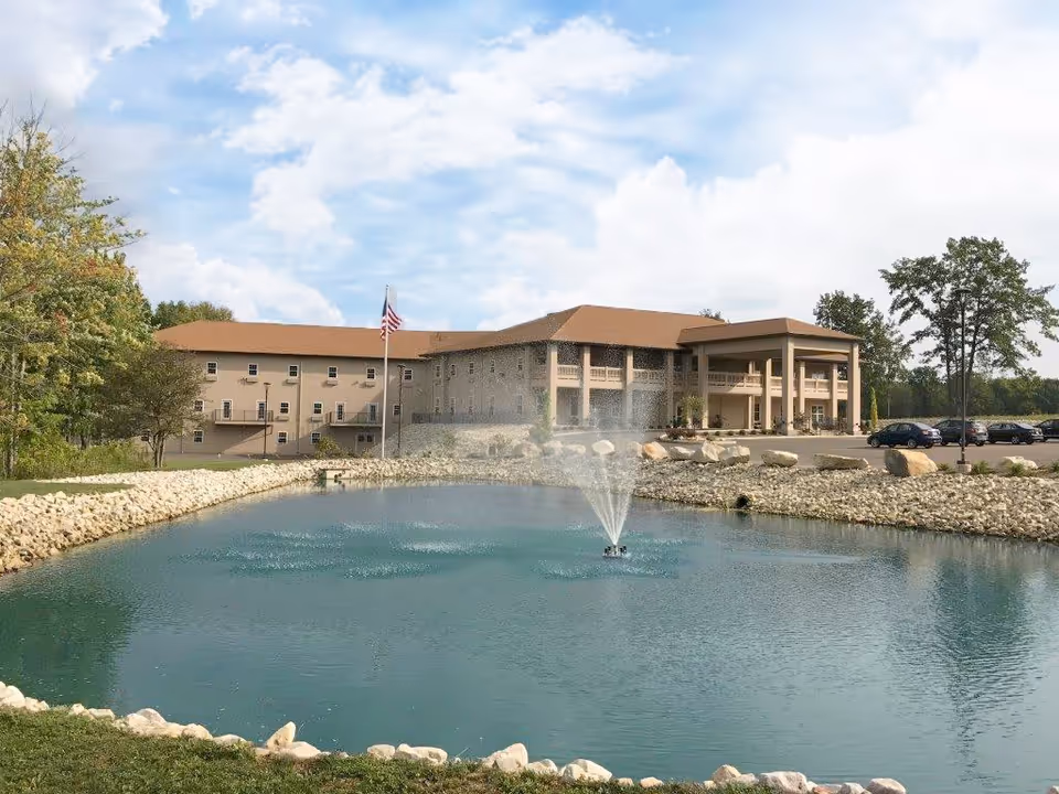 Exterior view of The Lakes at Jefferson senior living facility showing a large pond with a water fountain in the foreground, a beige multi-story building with a brown roof in the background, an American flag on a flagpole near the building, several parked cars, and trees surrounding the area under a partly cloudy sky.