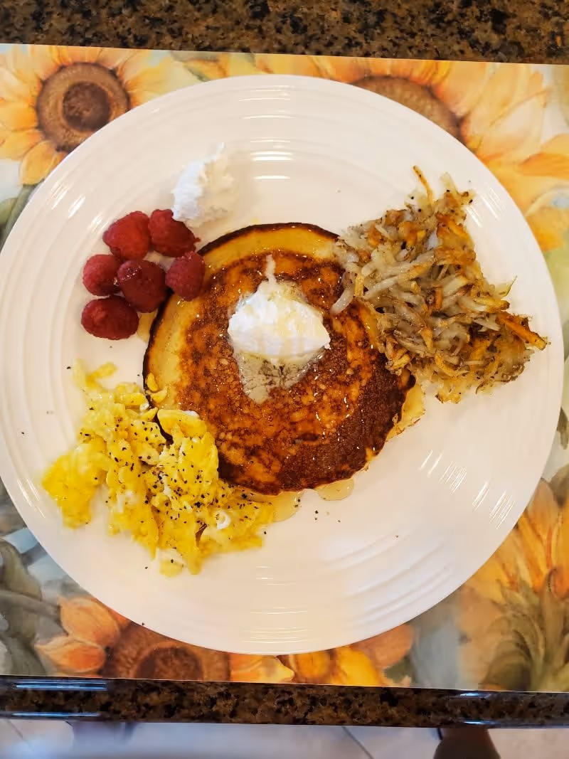 A white plate with a breakfast meal consisting of a pancake topped with butter and syrup, scrambled eggs with black pepper, a serving of hash browns, a dollop of whipped cream, and several raspberries. The plate is placed on a sunflower-themed placemat on a granite countertop.