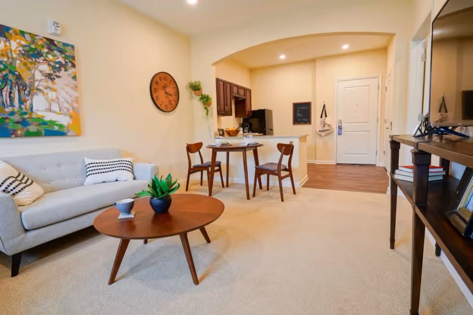 A cozy living area in a senior living facility featuring a light gray sofa with patterned pillows, a round wooden coffee table with a small plant and a cup, a small dining table with two chairs set with cups and plates, a wall clock, colorful abstract wall art, and a view into a compact kitchen and entrance door.
