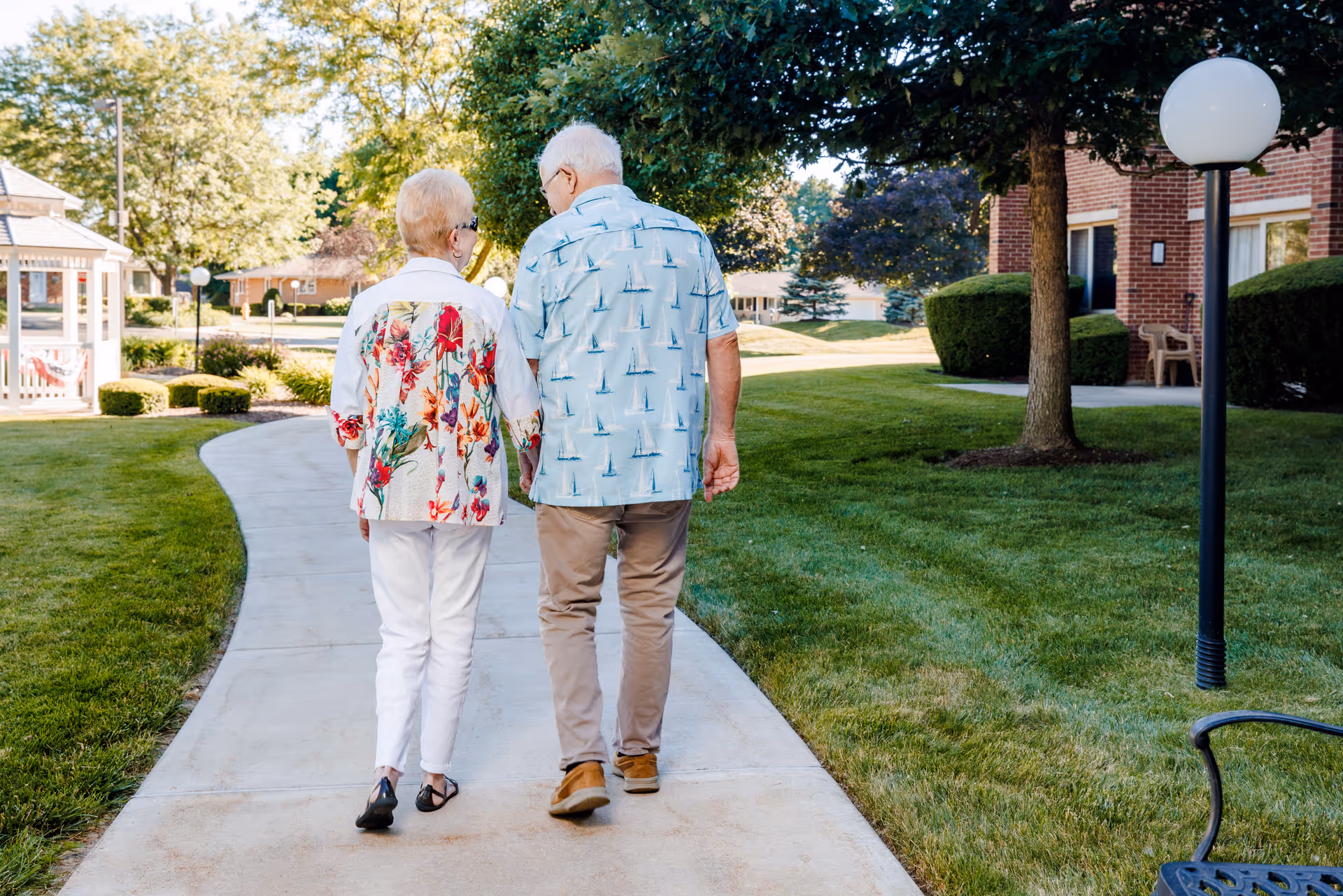 An elderly couple walking hand in hand on a paved pathway through a well-maintained garden area with green grass, trees, and a brick building in the background during daytime.