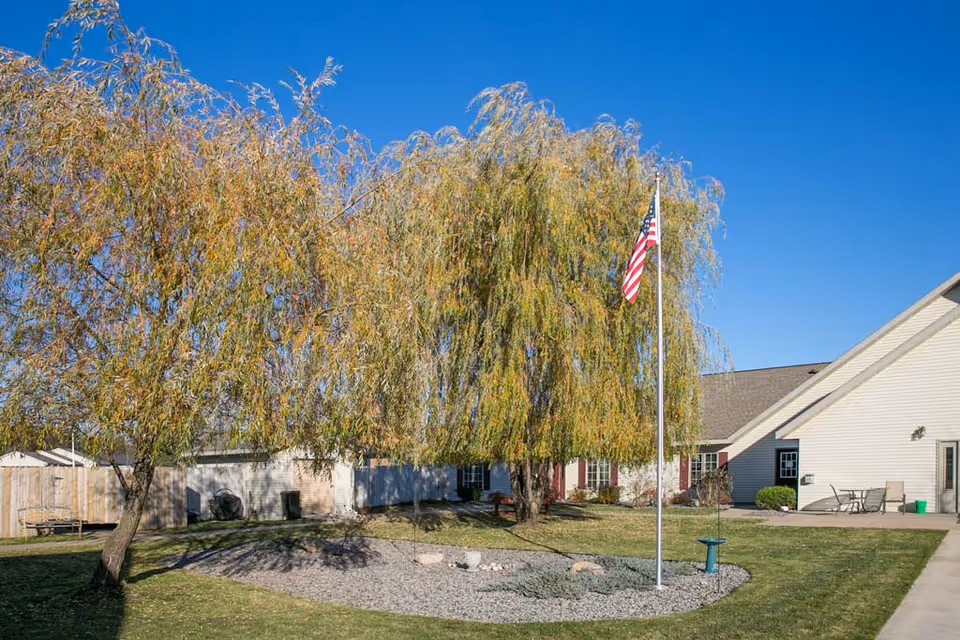 Outdoor area of Cranberry Court Assisted Living featuring a green lawn, two large willow trees, a flagpole with an American flag, a circular rock garden, and a beige building with a patio and outdoor furniture under a clear blue sky.
