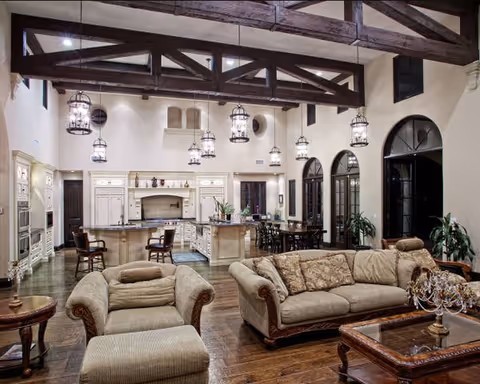 Open-concept living room and kitchen with exposed wooden beams, hanging lanterns, beige sofas, and hardwood floors.