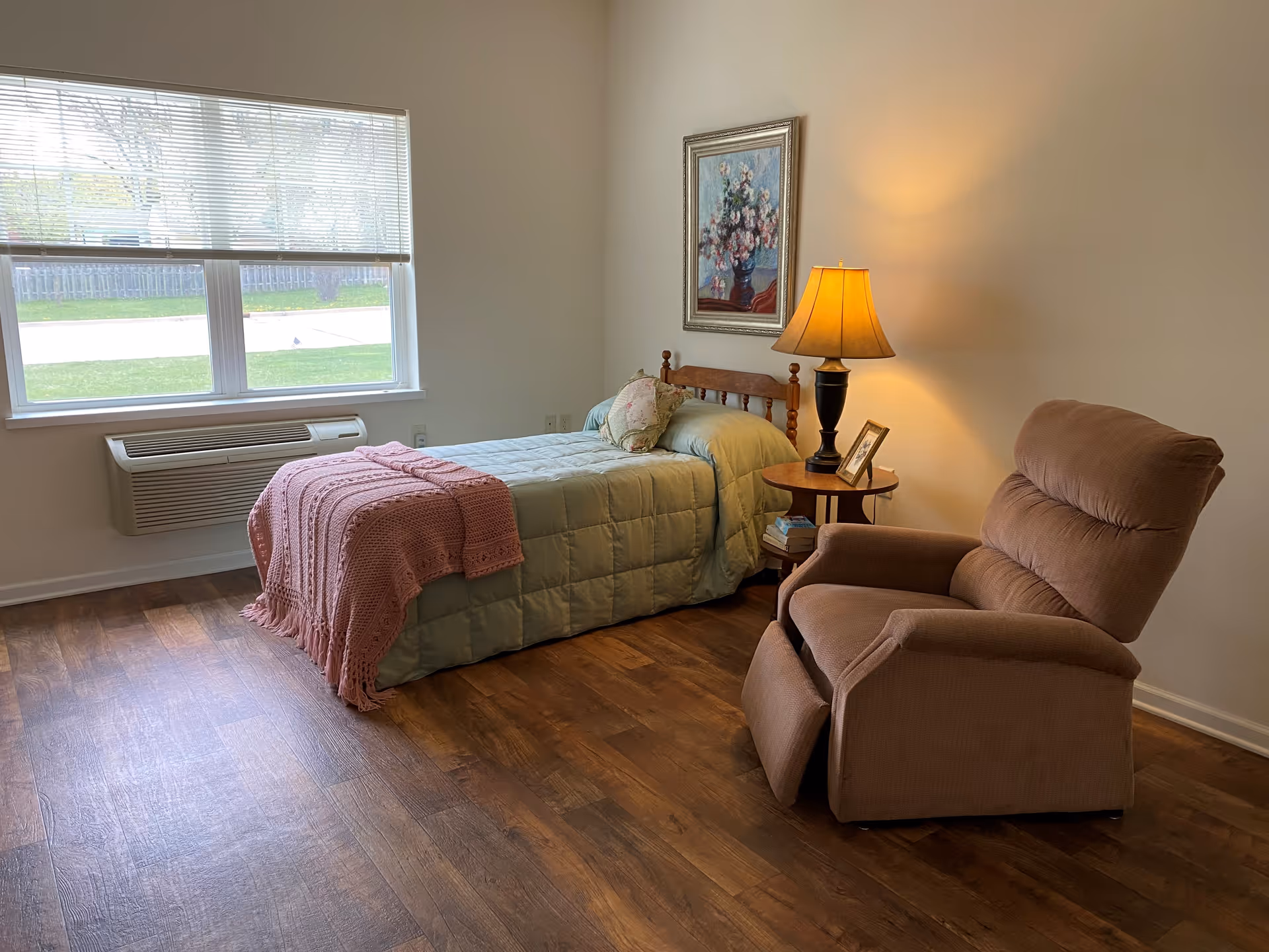 A cozy bedroom with a single bed covered in a green comforter and a pink knitted throw blanket. Next to the bed is a wooden side table with a lamp, a framed photo, and some books. A comfortable brown recliner chair is positioned nearby. The room has wooden flooring, a large window with blinds, and a framed floral painting on the wall.
