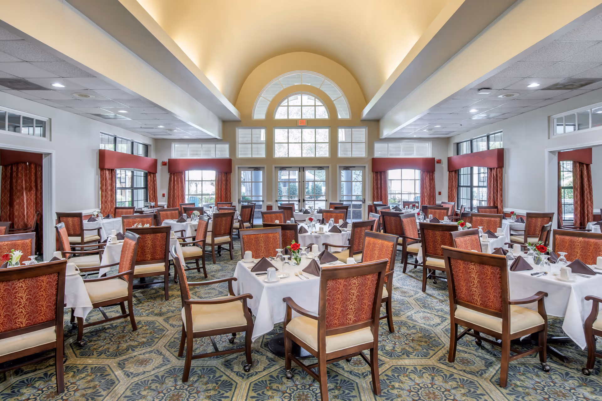 A spacious dining room with multiple tables covered in white tablecloths, each set with cups, glasses, silverware, and napkins. The chairs have wooden frames with red patterned upholstery. Large windows with red curtains allow natural light to fill the room, and an arched window above the double glass doors adds architectural interest. The carpet has a decorative pattern, and the ceiling is vaulted and painted a soft yellow.