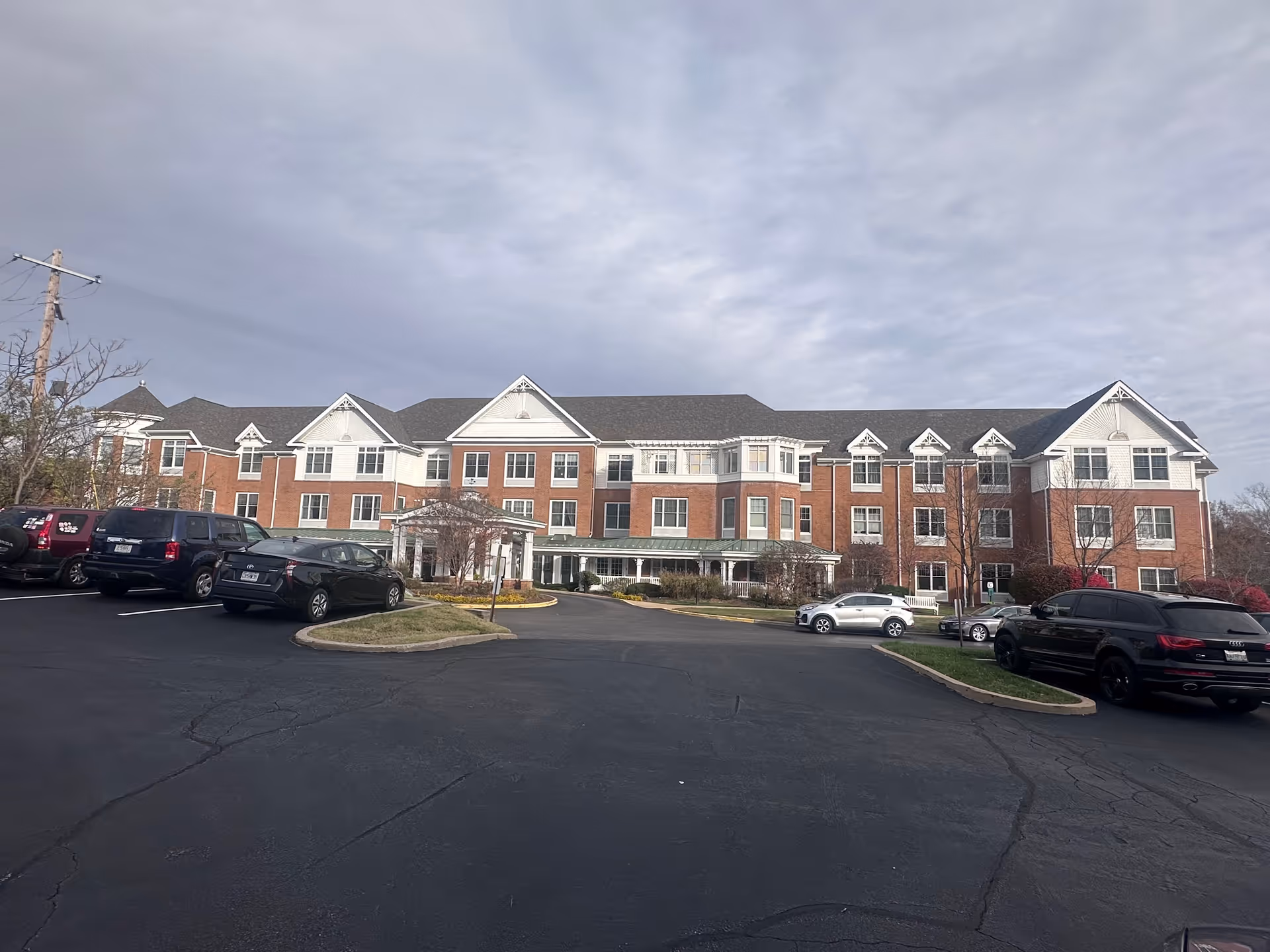 Front exterior view of a large senior living facility building with red brick and white siding, multiple windows, and a covered entrance. Several cars are parked in the paved parking lot in front of the building under a cloudy sky.