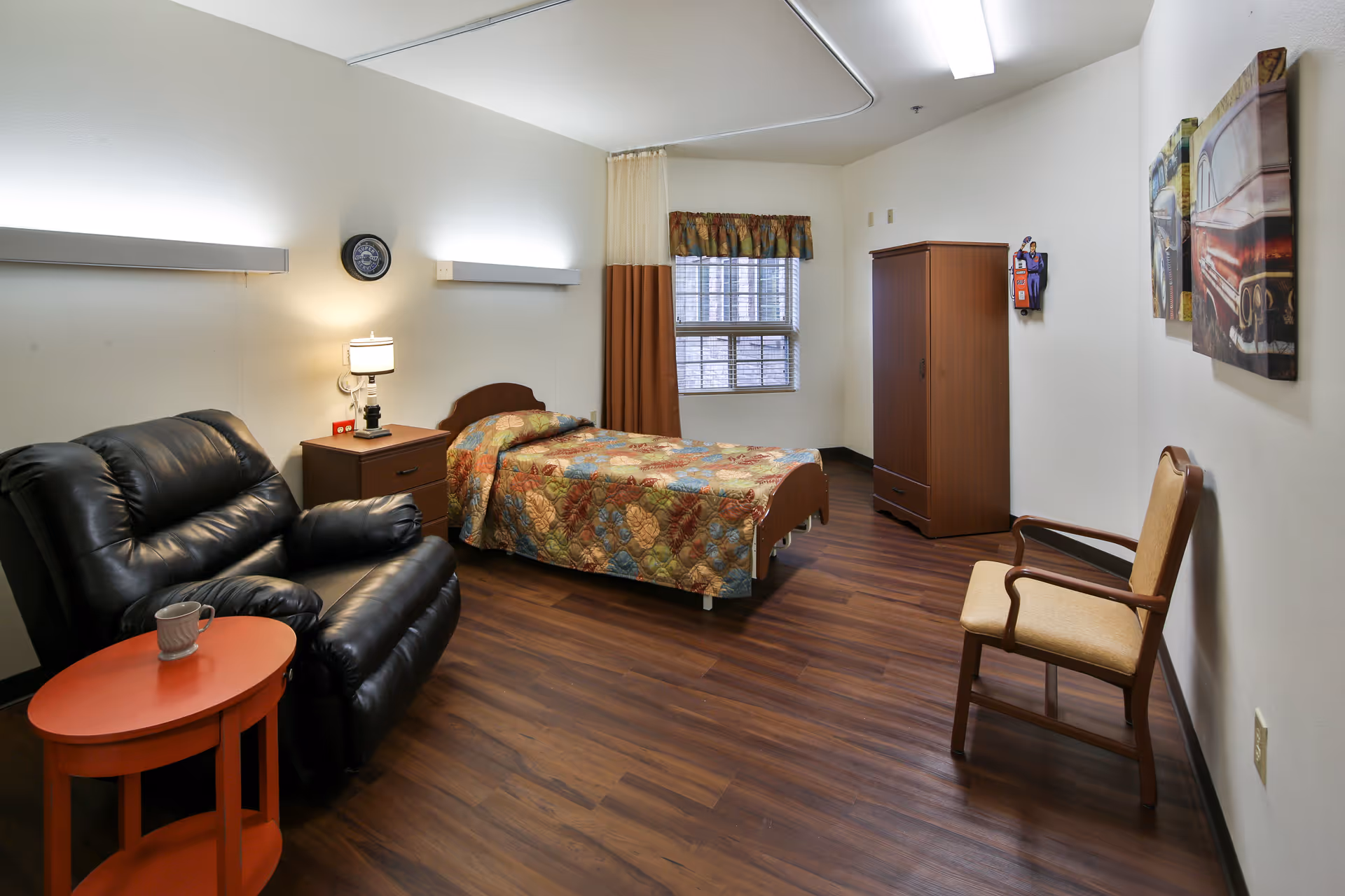 A senior living facility bedroom with a single bed covered in a colorful quilt, a wooden nightstand with a lamp, a black leather recliner chair next to a small round orange table with a cup on it, a wooden wardrobe, a beige armchair, and two framed pictures on the wall. The room has wood flooring, a window with curtains, and bright overhead lighting.