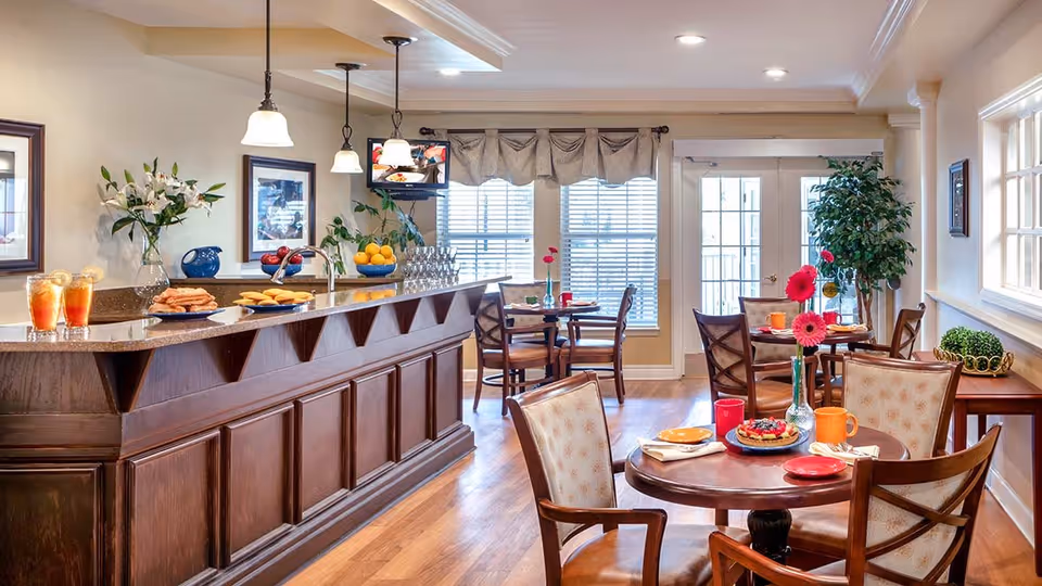 A bright and inviting dining area in an assisted living facility featuring a wooden bar counter with hanging pendant lights, plates of cookies and glasses of iced tea. Several round tables with chairs are set with colorful plates, cups, and vases holding pink flowers. Large windows with blinds and a glass door allow natural light to fill the room, complemented by indoor plants and framed artwork on the walls.